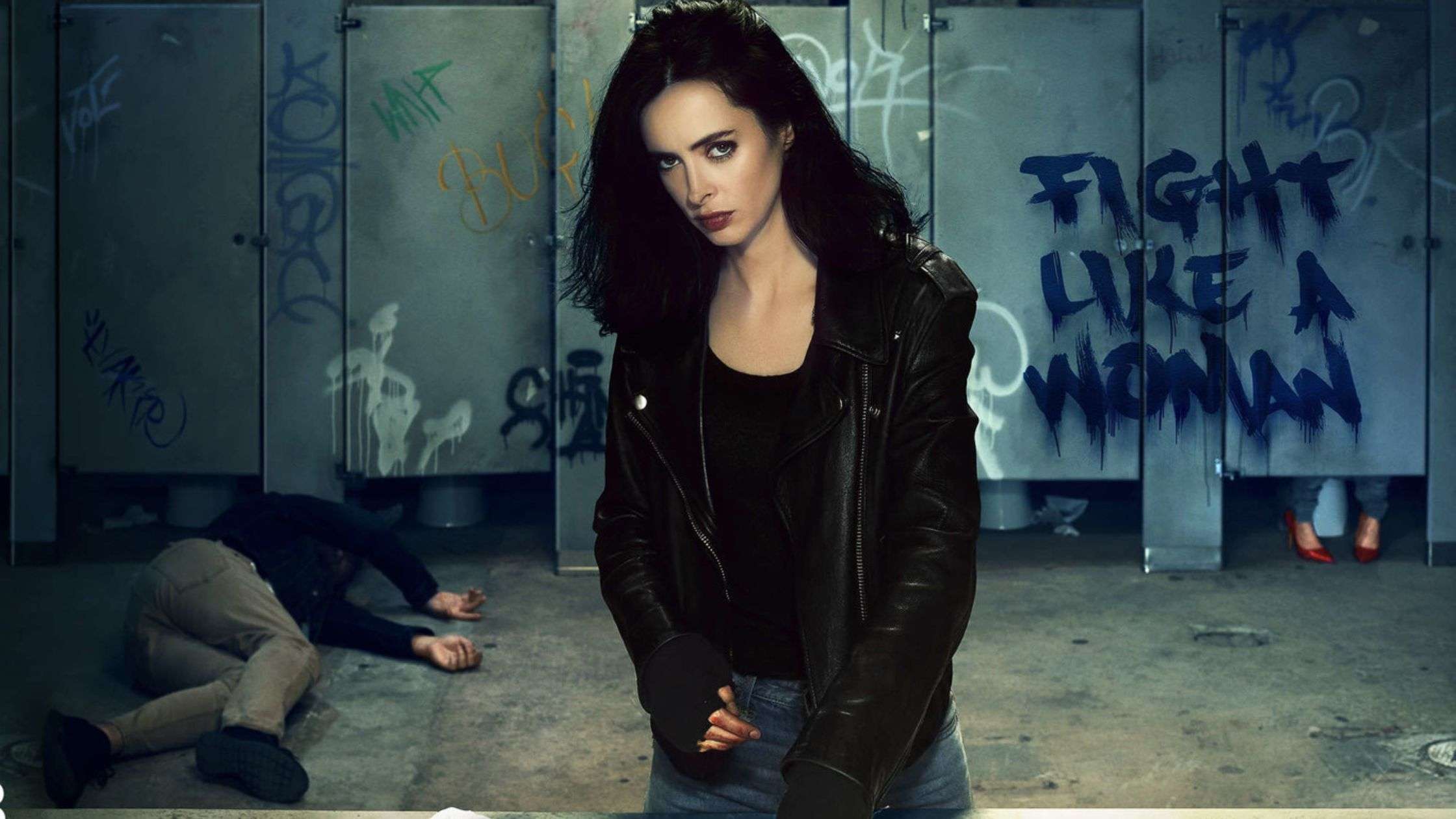 Jessica Jones con chaqueta de cuero en un baño lleno de grafitis, mensaje Fight like a woman en la pared