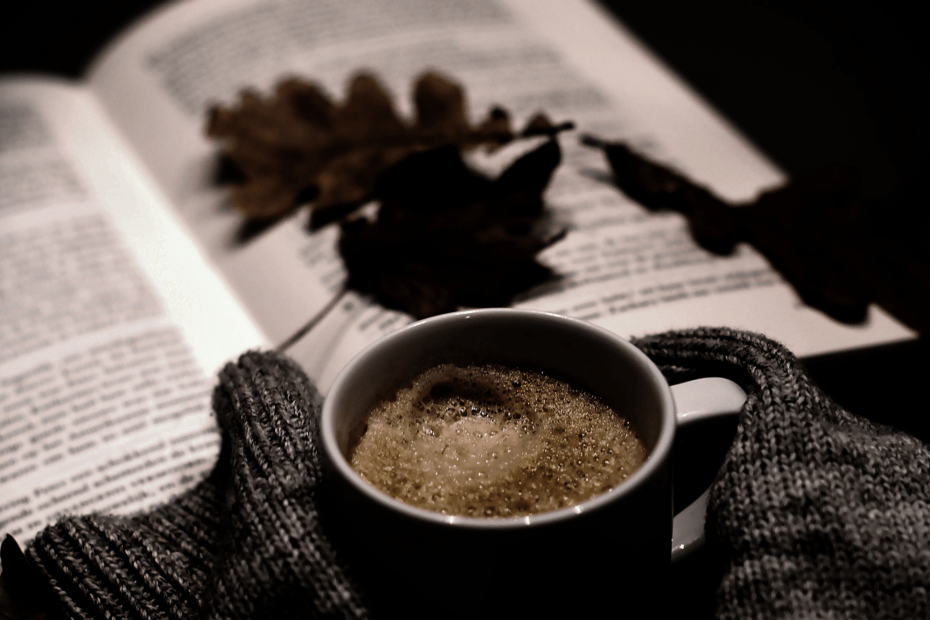 Taza de café entre guantes de lana sobre un libro abierto con hojas secas, en penumbra. Imagen que refleja la mezcla de calidez y misterio del cozy crime oscuro.