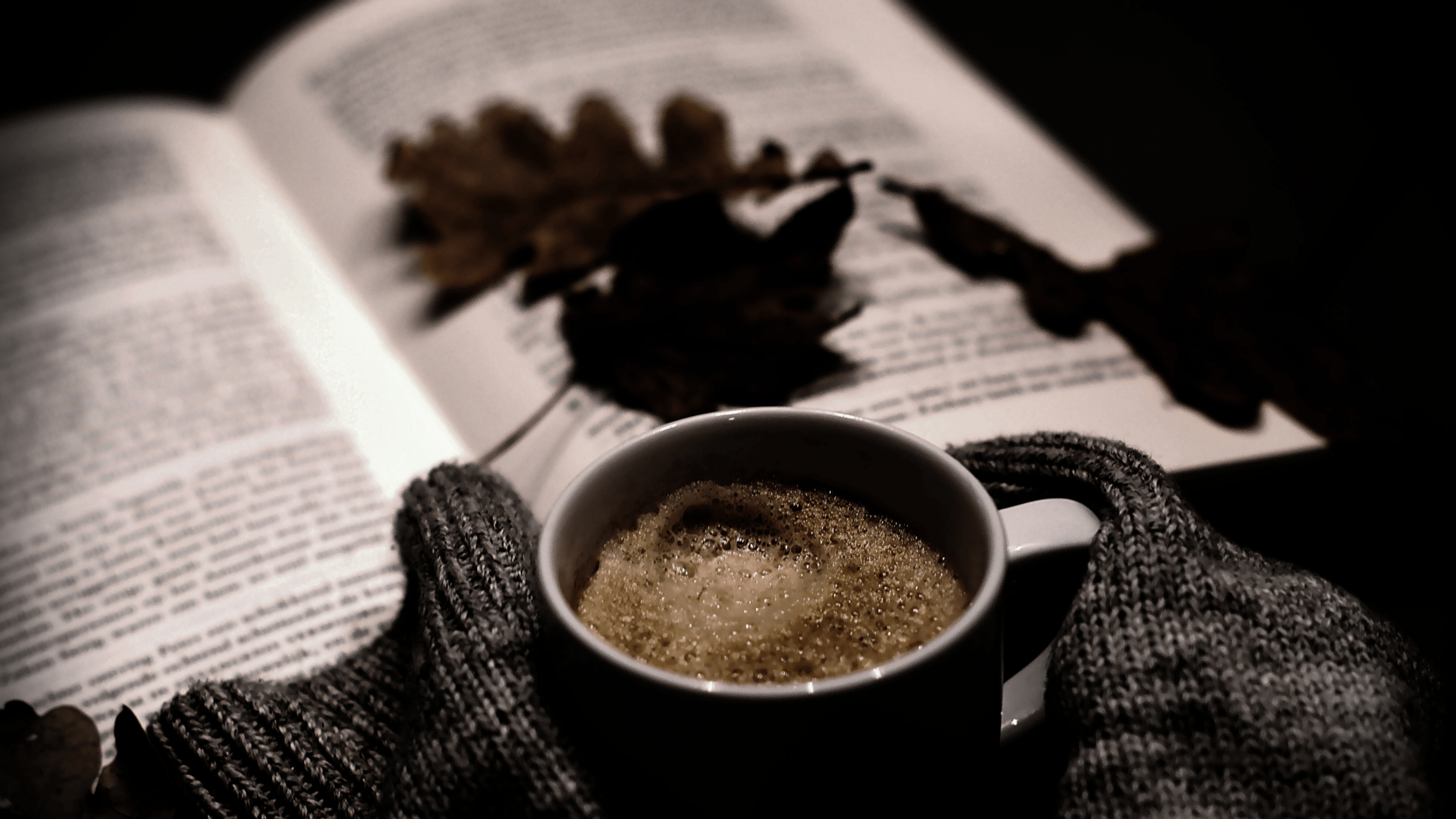 Taza de café entre guantes de lana sobre un libro abierto con hojas secas, en penumbra. Imagen que refleja la mezcla de calidez y misterio del cozy crime oscuro.