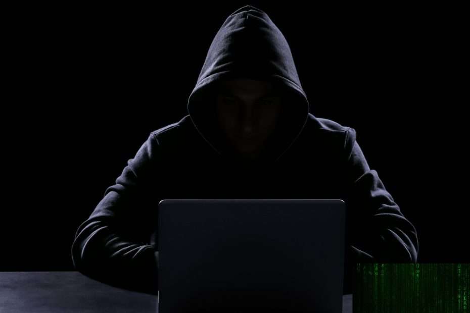 Silueta oscura de un hacker con capucha frente a un ordenador portátil, representando la figura del 'asesino invisible' en la novela negra contemporánea y el peligro del crimen digital.